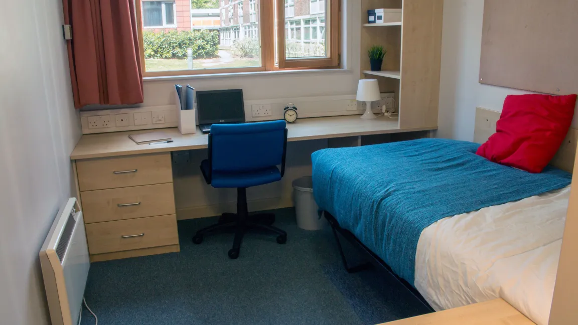 An empty student bedroom containing a neatly made single bed and a large desk with chair, beneath a window.