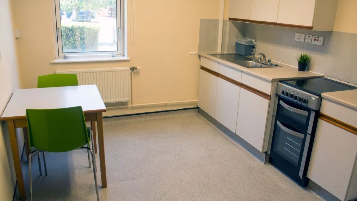 A large kitchen with laminate flooring. It contains modern looking kitchen appliances and cupboards, a small square table with two chairs and a window.