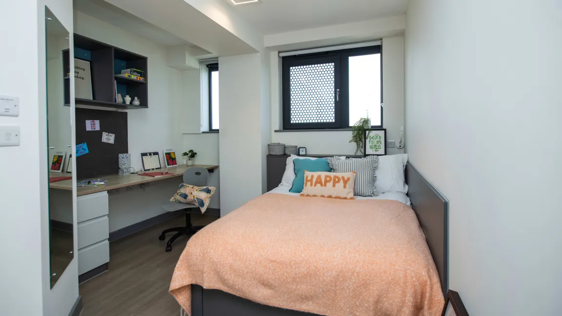 A standard bedroom showing a bed, a desk with shelves above and a chair.
