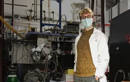 Engineering student Oliver Hitchens in a lab coat and mask in front of lab equipment