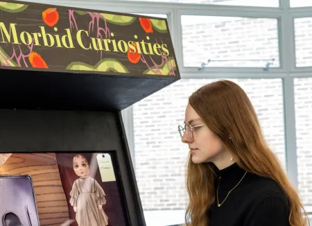 Laura Alford, BA Games Design and Art student looking at an arcade game console named Morbid Curiosities. There is a child like doll on the screen and 2 candle sticks on the game console. 