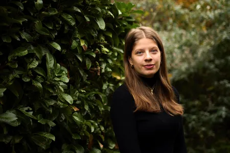 Head and shoulders portrait image of Paulina Borek, MA History student smiling and looking at the camera. She is outside, standing in front of dark green shrubs.