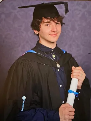 Samual Marsh dressed in his graduation robes and mortar board, holding a paper degree rolled up and tied with a blue ribbon.