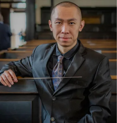 Xinda Zhu is sitting in an auditorium, looking at the camera and holding a baton.