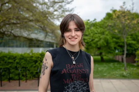 A student stands smiling outside the Physics building dressed casually in a sleeveless band T-shirt.