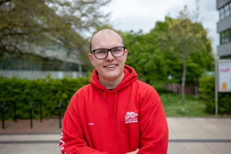 A headshot of a student on a university campus. They are wearing a red hoodie and glasses.