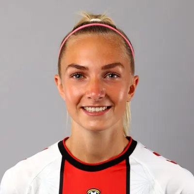 Pheobe Williams smiles at the camera. She is wearing her teams football shirt and has her blonde hair tied back, held out of her eyes with a headband