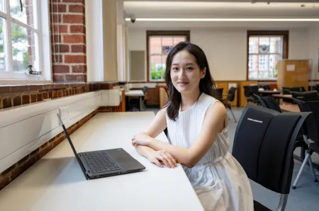 Lin sitting at a desk in an empty classroom. She has a laptop on the desk in front of her, and she's smiling for the camera.
