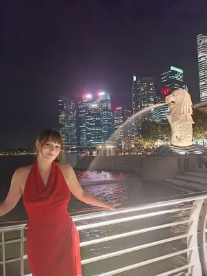 female student stood in front of railings with water feature and city skyline with lit up tower buildings against dark night sky in the background.