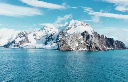 Snow-covered Antarctic island rising from clear blue ocean beneath a bright sky, with a small boat visible in the distance.