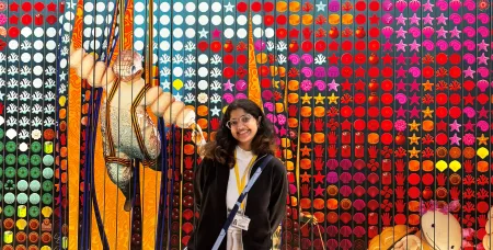Design Management student smiling in front of a colourful textile and craft exhibition display.