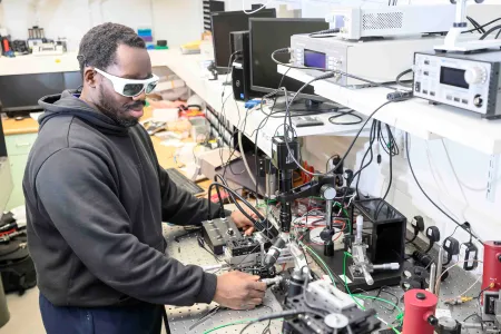 Student adjusts photonics laboratory equipment while wearing protective laser safety glasses.