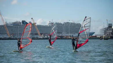 Three people windsurfing beneath blue skies on a sunny day. A large cruise ship is visible behind them, docked in port.