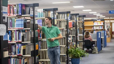 A student selects a book from a shelf while other students work at desks in the background