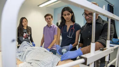 A group of students watch as one of them works on a dummy patient in a hospital bed