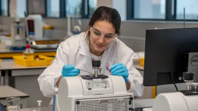 A student in a lab coat uses a metal spatula to collect a small sample specimen from a piece of technical equipment. Behind her, a large modern laboratory full of equipment and glassware can be seen.