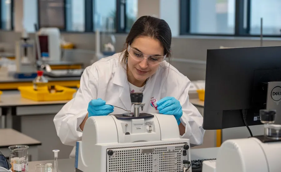 A student in a lab coat uses a metal spatula to collect a small sample specimen from a piece of technical equipment. Behind her, a large modern laboratory full of equipment and glassware can be seen.