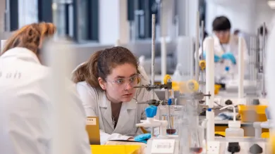 A student in a lab coat crouches down to closely monitor a chemistry experiment being conducted in a large piece of glassware.