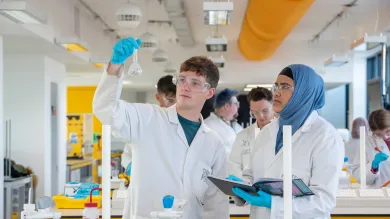 Two students wearing lab coats and protective glasses conduct an experiment. One holds up a glass beaker, while the other takes notes. They are in a laboratory, surrounded by a variety of chemistry equipment.