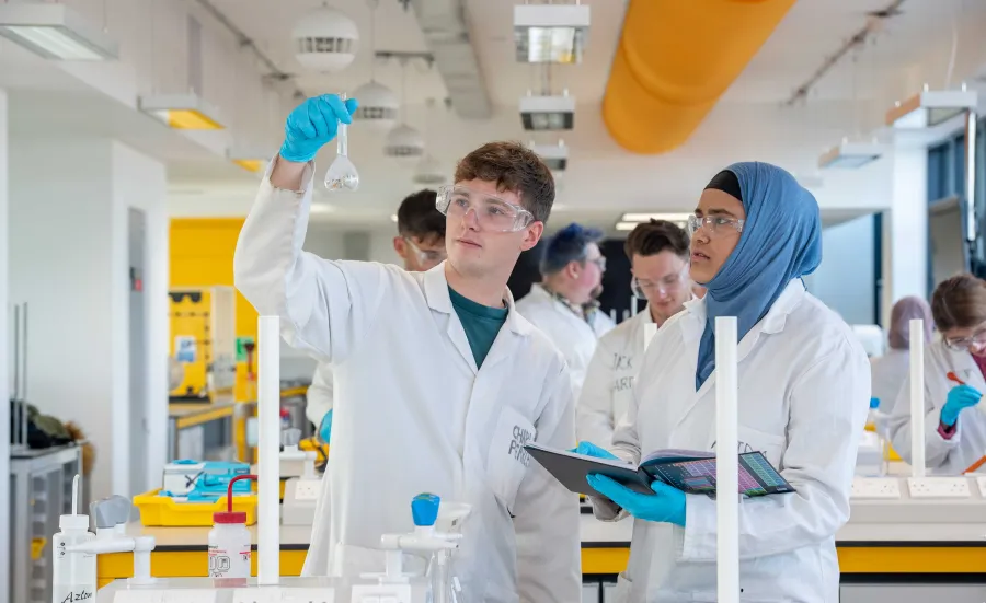 Two students wearing lab coats and protective glasses conduct an experiment. One holds up a glass beaker, while the other takes notes. They are in a laboratory, surrounded by a variety of chemistry equipment.