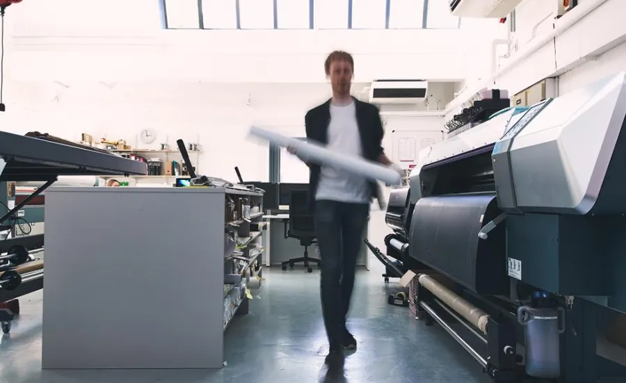 A student walking between several industrial-sized printers and a long work bench lined with guillotine paper cutters