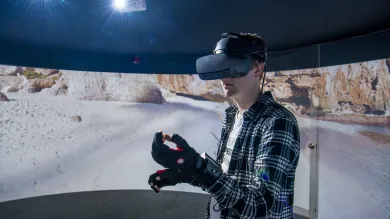 A student wearing a virtual reality headset and gloves, standing in front of a 360 degree video wall displaying what he can see.