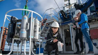 Three students outside on the deck of a boat, on a sunny day. One crouches down to collect a water sample from a large piece of technical equipment.