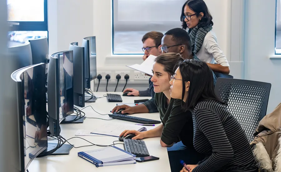 Several students working together across a row of workstations with curved monitors.