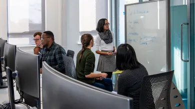A group of students discussing their notes on a whiteboard in the conference room, as 2 fellow students consult their work on a computer behind them.