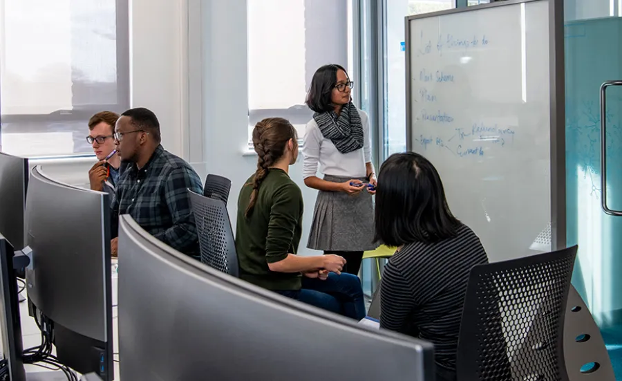 A group of students discussing their notes on a whiteboard in the conference room, as 2 fellow students consult their work on a computer behind them.