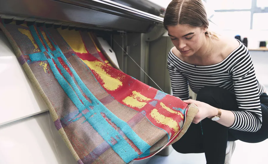 A student crouches by a computerised flat knitting machine as her creation works its way through the mechanism.