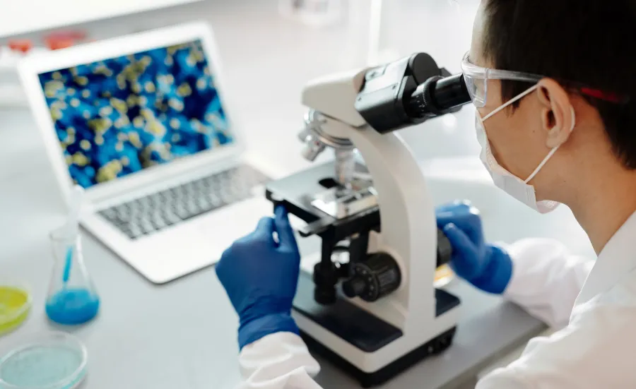 Student in a lab coat is looking into a microscope and showing results on the laptop infront of her