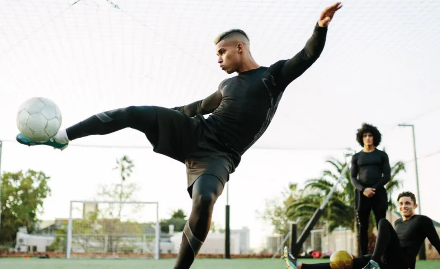 A young male footballer raising his leg parallel to the ground, to kick a ball towards the camera.