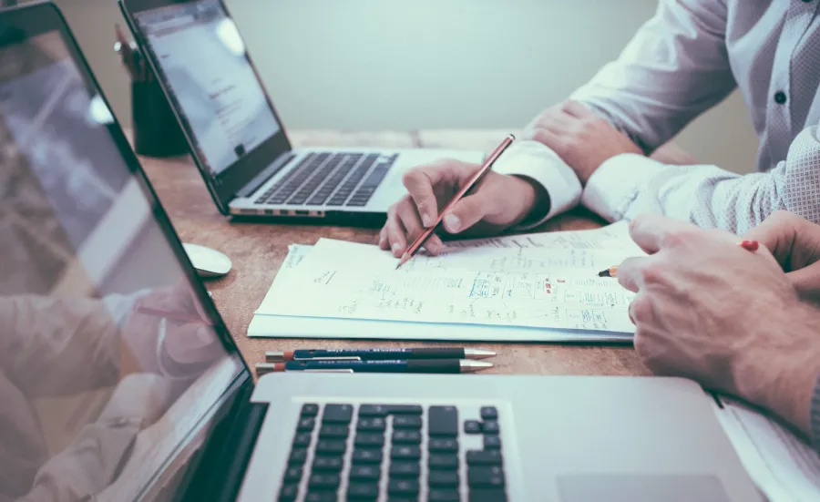 Close up view of two people working at a desk with laptops, paper and pens