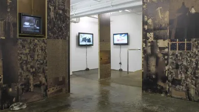 The Taplow House art installation in ASC Gallery. The installation walls in the foreground are covered in dark, industrial images. In the background, they are white. Several televisions hang on the walls.