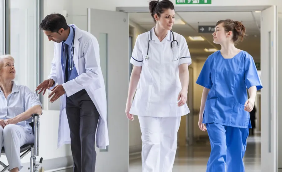 A wide shot of a hallway at Southampton General Hospital. To the left, a doctor is speaking to a patient in a wheelchair. To the right, a doctor and medical student are walking as they talk.