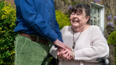 A woman in late middle age sits and holds hands of a man standing next to her