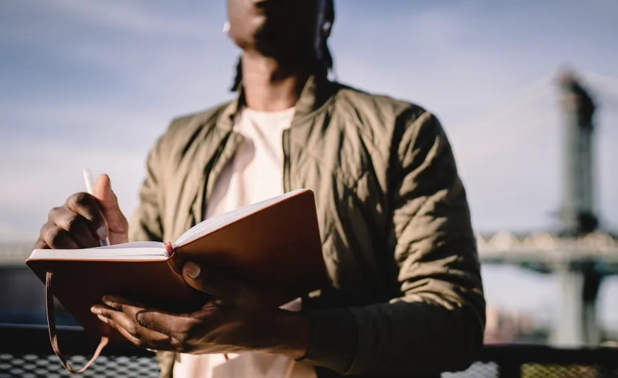 A Black man writes on a notepad as he stands outside, a blue sky and a bridge behind 