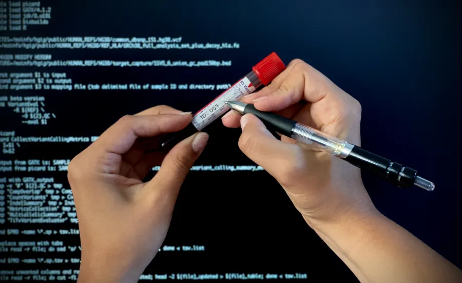 Close-up of hands labelling a patient blood sample for molecular analysis. In the background a computer screen shows programme code to process sequencing data. 