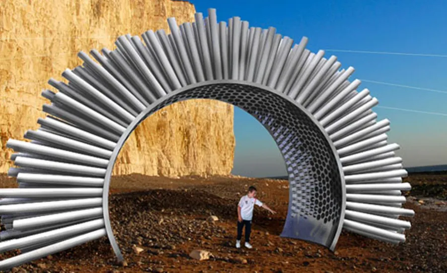 A student stands beneath a long arch with metal tubes extending outwards across its length. The student is pointing up at this acoustic device, which is on a beach