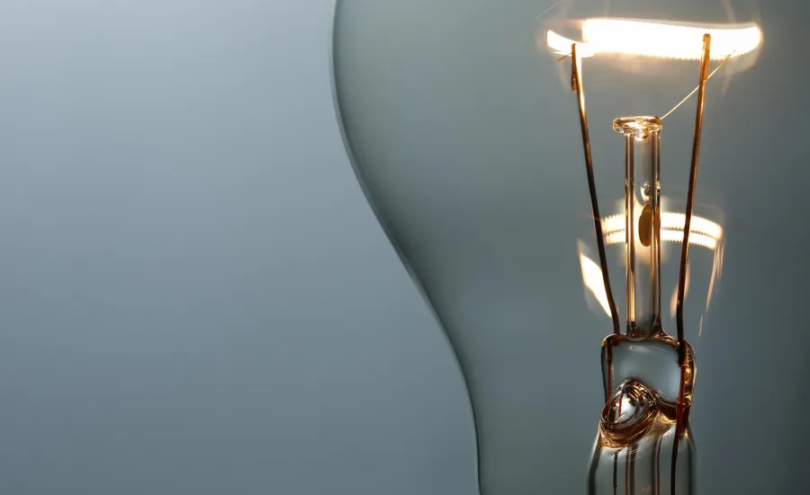 A close up of the inside of a light bulb focuses on the filament of tungsten wire that electricity runs through, a warm, lit colour in contrast to the grey background. Light bulbs are sometimes used as a way to communicate that someone's had a good idea or some insight.