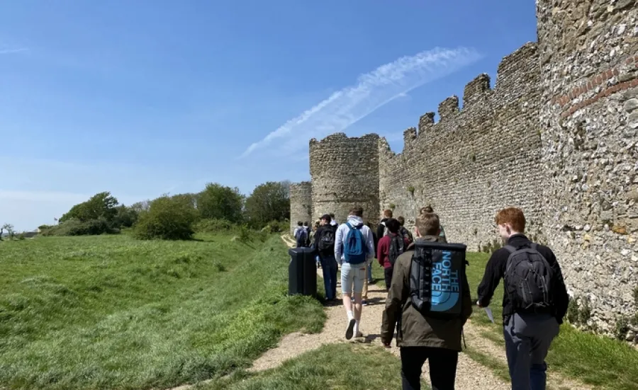 A group of students walks away from us, by a historic castle. The two closest to us are in discussion, beneath the wall that stretches away from them, to a tower of the castle and, perhaps, the castle entrance. In the foreground, we can make out each individual stone that makes up this structure.