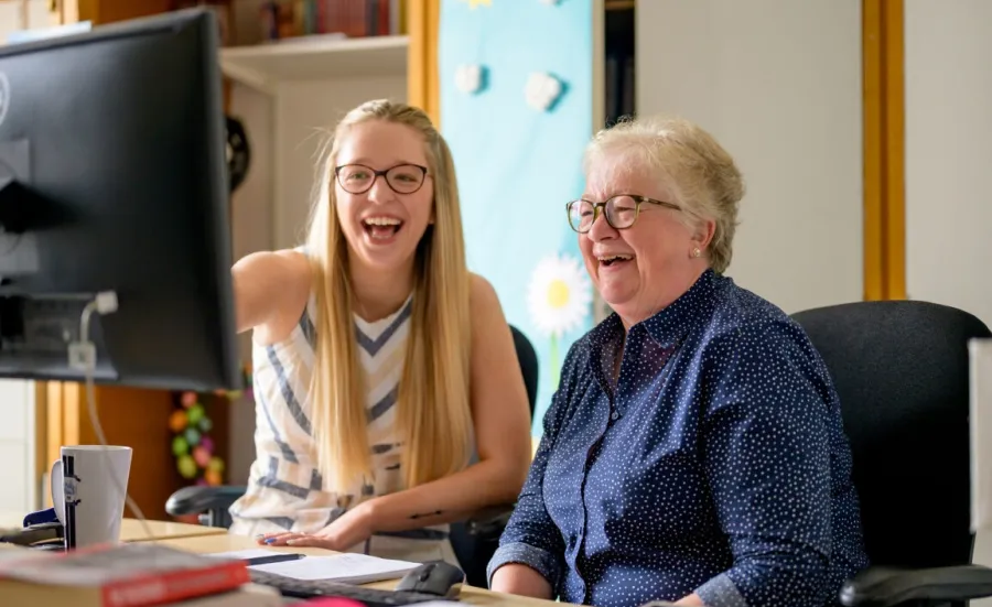 Two laughing women, one young, one older, sit in front of a computer screen. The young woman points at the screen.