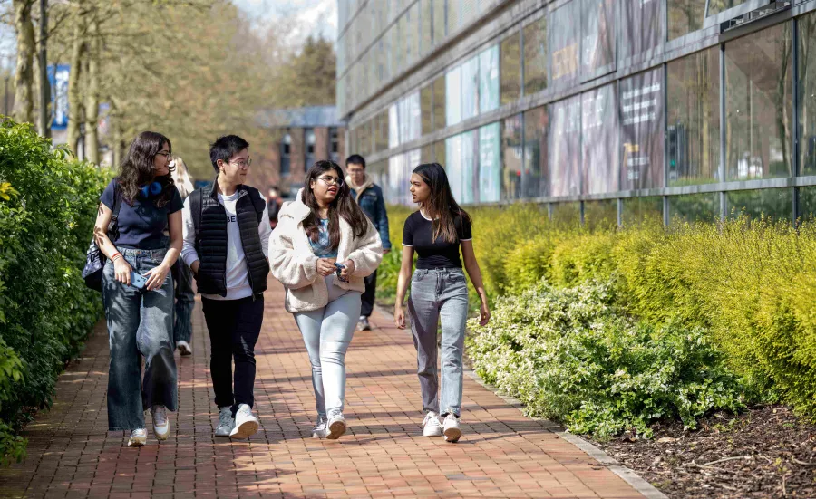 A group of students of different ethnicities walk through a green, sunlit university campus, turned towards each other, in discussion