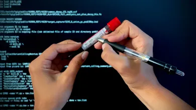 Somebody writes the time and date on a blood sample in a vial, against a background of a screen of data to represent genomic data. 
