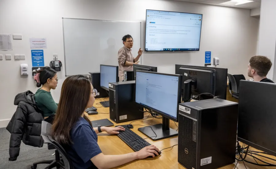 Three students in front of monitors in a classroom listen as a lecturer makes a point