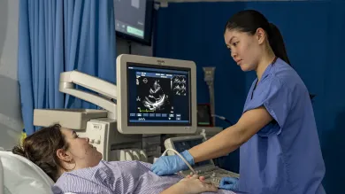 Healthcare students training in a hospital setting. One student poses as a patient, the other uses monitoring equipment with a scanner and screen