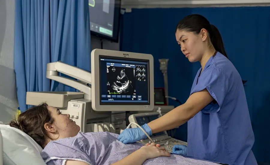 Healthcare students training in a hospital setting. One student poses as a patient, the other uses monitoring equipment with a scanner and screen