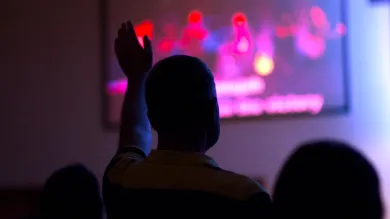 People in a room silhouetted against a screen. The image on the screen is unclear. One of the people has their arm raised to ask a question.
