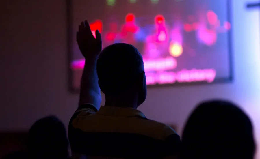 People in a room silhouetted against a screen. The image on the screen is unclear. One of the people has their arm raised to ask a question.